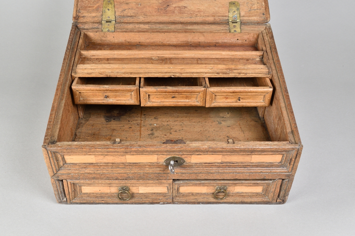 Skrin tillverkat i ek, med rik intarsia av två andra träslag. Rektangulär till formen med pulpetlock. Locket med profilerad och centrerad spegel. Framsida med nyckelhål och nyckelskylt, samt två lådor med draghandtag av mässing. Lådorna går att låsa från skrinets insida med hjälp av två träpluggar. Insidan av skåpet försedd med avsats för placering av skrivredskap samt tre mindre lådor med dragknoppar. 