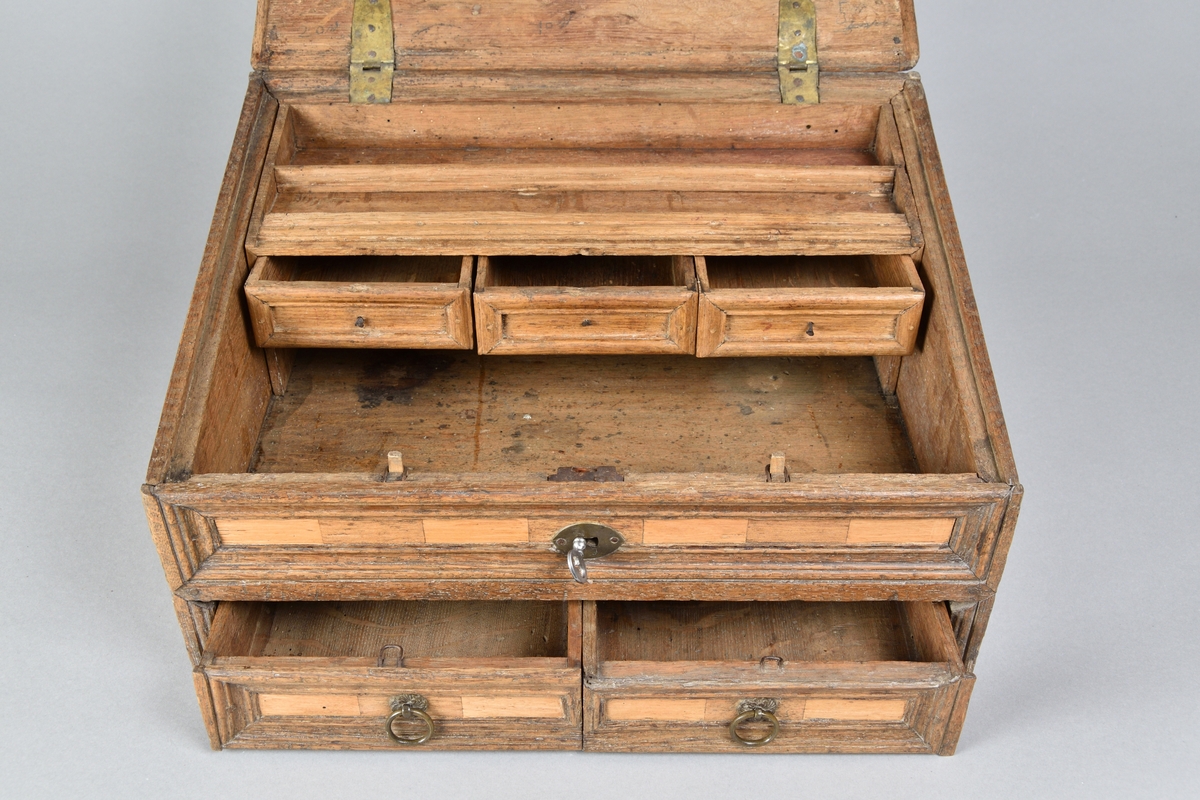 Skrin tillverkat i ek, med rik intarsia av två andra träslag. Rektangulär till formen med pulpetlock. Locket med profilerad och centrerad spegel. Framsida med nyckelhål och nyckelskylt, samt två lådor med draghandtag av mässing. Lådorna går att låsa från skrinets insida med hjälp av två träpluggar. Insidan av skåpet försedd med avsats för placering av skrivredskap samt tre mindre lådor med dragknoppar. 