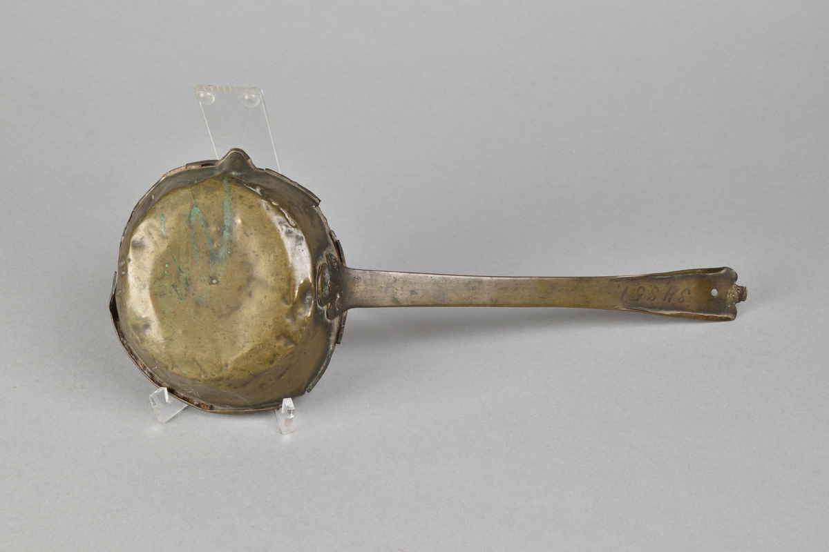 Skopa tillverkad i mässing. Cirkelrund skopa med snip och rullad överkant. Handtag fäst med nitar. Med graverad ornering i form av en krona över initialerna "RSE", samt graverat årtal "1677" och initialerna "I D".
