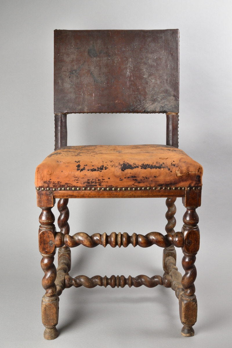 Stol tillverkad av björk, i barockstil. Ryggbricka, ryggstolpar och sits klädda i skinn, fäst med nitar. Fyra raka ben, med stabiliserande framslå, samt ett stabiliserande H-format benkryss. Ben och slår delvis profilsvarvade och delvis spiralsvarvade. Benen med konformade fotavslut. 