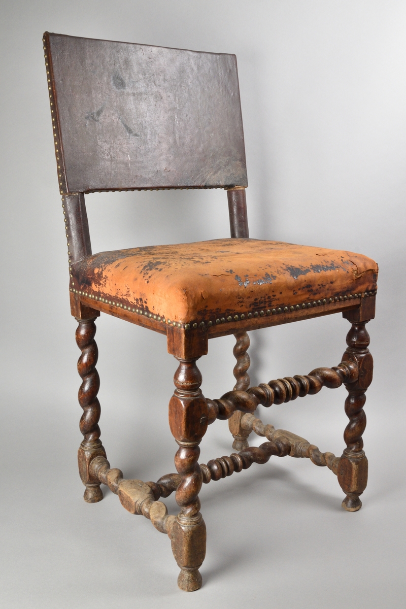 Stol tillverkad av björk, i barockstil. Ryggbricka, ryggstolpar och sits klädda i skinn, fäst med nitar. Fyra raka ben, med stabiliserande framslå, samt ett stabiliserande H-format benkryss. Ben och slår delvis profilsvarvade och delvis spiralsvarvade. Benen med konformade fotavslut. 