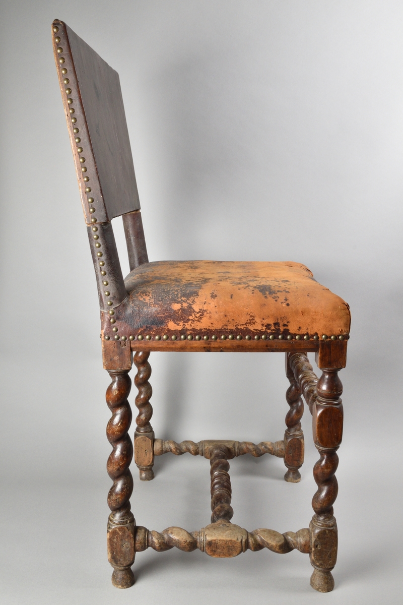 Stol tillverkad av björk, i barockstil. Ryggbricka, ryggstolpar och sits klädda i skinn, fäst med nitar. Fyra raka ben, med stabiliserande framslå, samt ett stabiliserande H-format benkryss. Ben och slår delvis profilsvarvade och delvis spiralsvarvade. Benen med konformade fotavslut. 