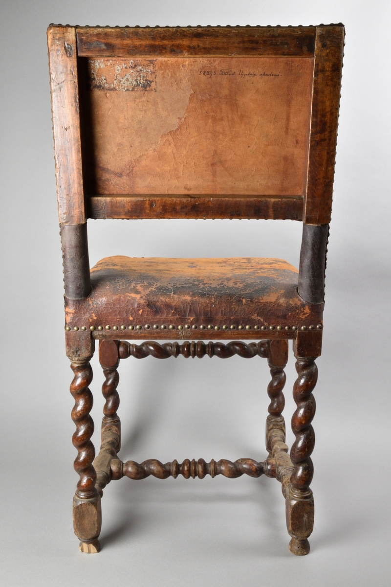 Stol tillverkad av björk, i barockstil. Ryggbricka, ryggstolpar och sits klädda i skinn, fäst med nitar. Fyra raka ben, med stabiliserande framslå, samt ett stabiliserande H-format benkryss. Ben och slår delvis profilsvarvade och delvis spiralsvarvade. Benen med konformade fotavslut. 