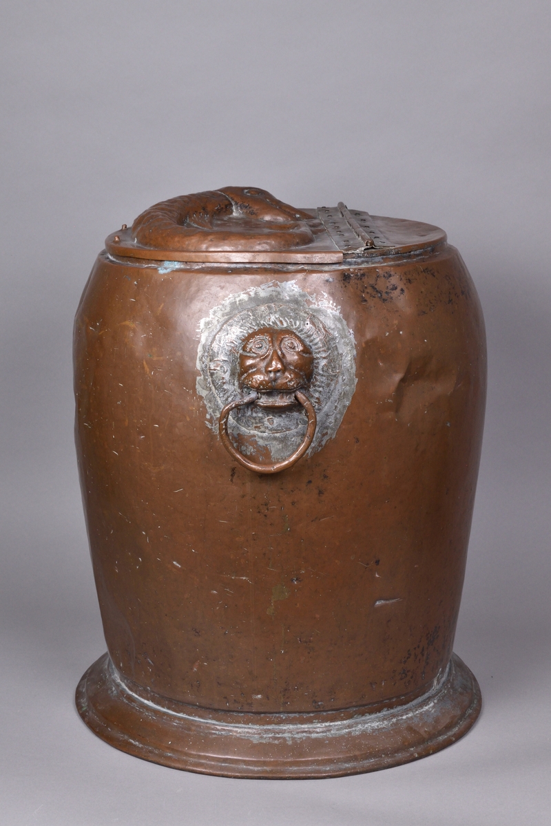 Kärl tillverkad av koppar. Hög och bred, med bukig överdel och avsmalnande underdel. Rakt avslut upptill med fällock på gångjärn, ornerat med driven dekor i form av fisk. Två handtag på var sida om kärlet, utformade som lejonhuvuden med ring. Kärlet vilar på en bred fotring.