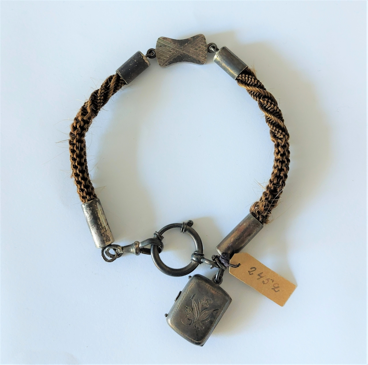 Urkedja av hår med silverbeslag. Med fyrkantig berlock med ingraverade blommor på ena sidan och en sköld på en den andra. Med en platta med ingraverat ”Minne”. - Stämpel B 7 = 1904, W. övriga stämplar oläsbara. -