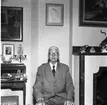 John Roman 70 år 1960. En äldre herre sitter i en hemmiljö och tittar in i kameran. På sin högra sida syns en sekretär och på hans vänstra en öppen spis. Det hänger tavlor på väggarna. Herren bär glasögon, kavaj och slips.