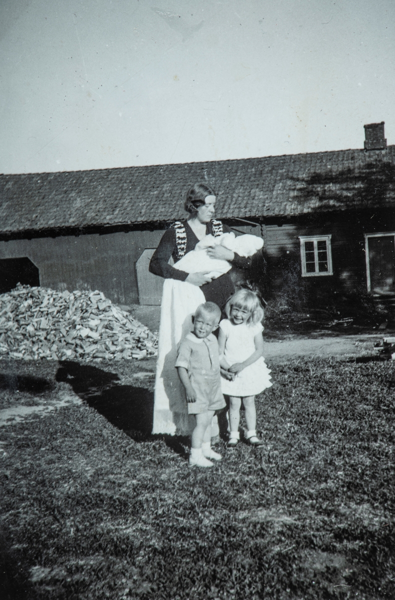 Øvre Tomter (60/1). Ingolf Prestruds barnedåp i 1935.  Fadder er Ingeborg Lunde på Ring f. Karset med dåpsbarnet. Dåpsbarnets søsken Helge Andreas og Helga står foran. Drengestua med nykløvd vedhaug i bakgrunnen.