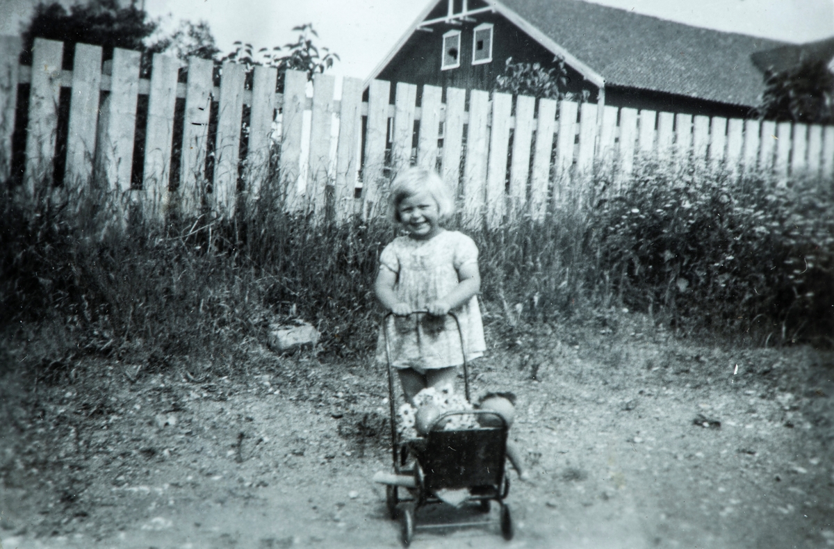 Øvre Tomter (60/1). Ragnhild Prestrud med dukkevogn. Stakitt mot hagen. Låven i bakgrunnen.