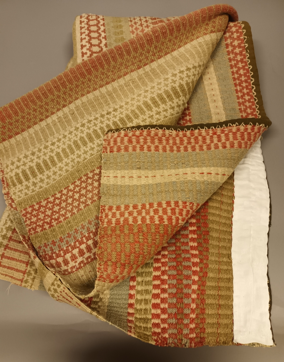 Krokbragdteppe kanta med ullstoff i kortendane. enning i bommull. Innslag av ullgarn. Plantefarga garn i innslag. Skjøyt p midten, bordane stemmer godt overens i skøyten.