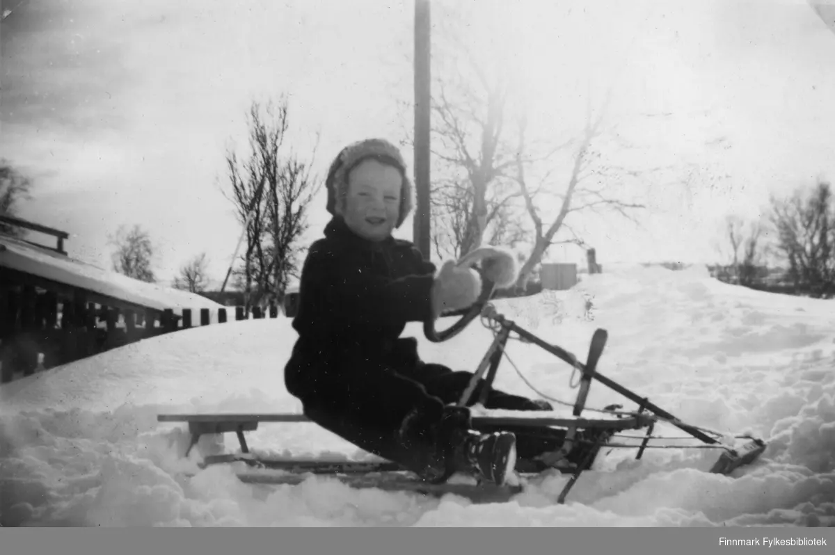 Bilde fra 1957 av meg sittende på en rattkjelke utenfor bakeriet i Varangerbotn.