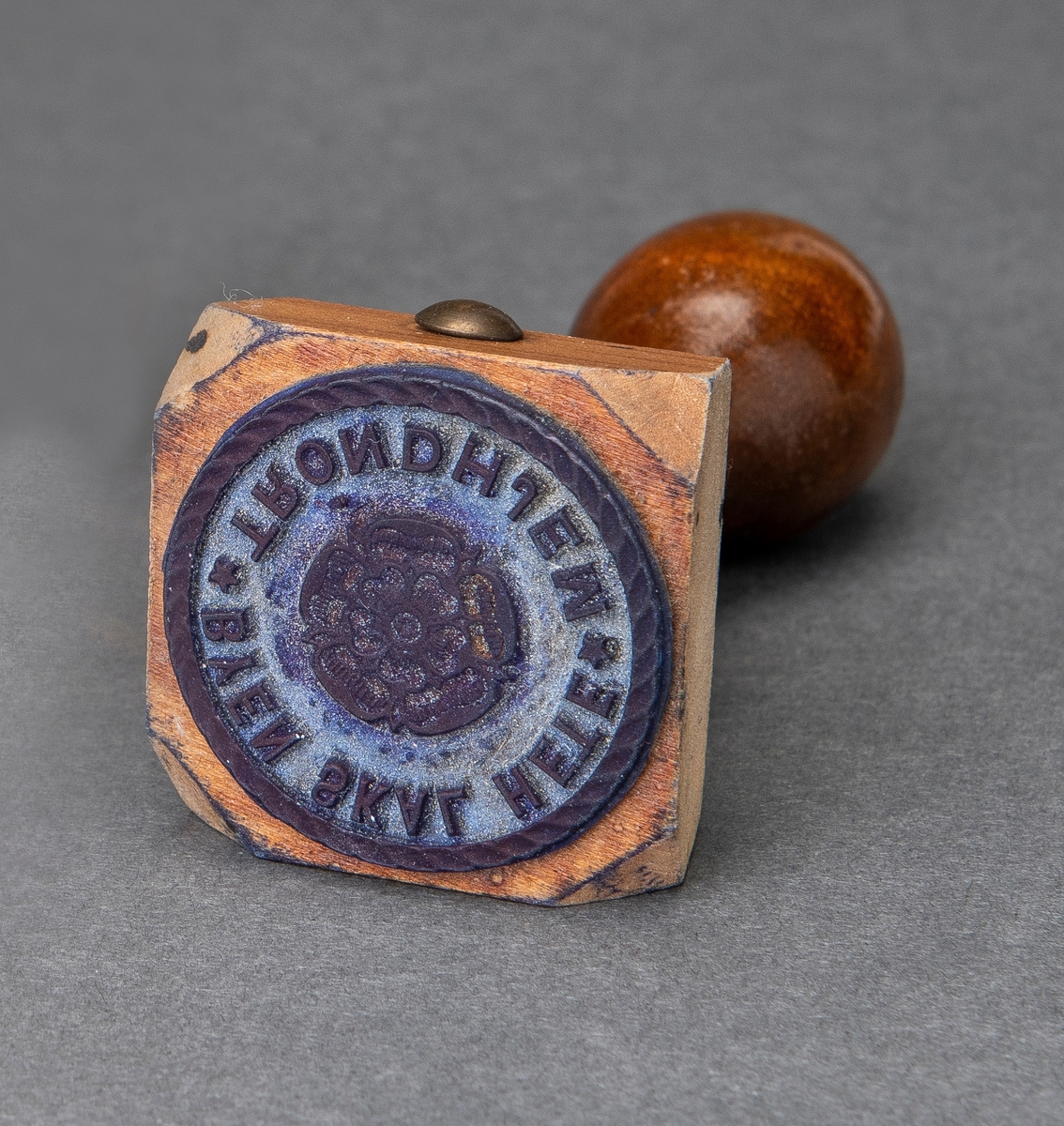 Stempel i lakkert tre. Selve stempelet har "Trondhjemsrosen" i midten omringet av teksten "Byen skal hete Trondhjem", slagord for striden om byen Trondheim sitt navn i årene 1929 og 1930.