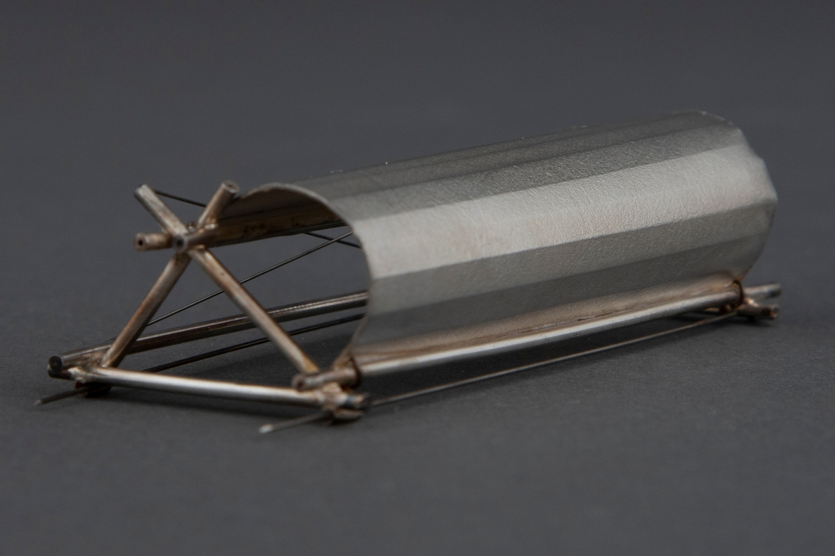 Brosje/smykkeobjekt formet av en rektangulær sølvplate som en halv sylinder med langsgående knekk. Denne er påsatt tynne sølvstrenger til en tilnærmet trekantform. Videre er det påsatt tynne ståltråder til dekor og som nåler til å feste brosjen.