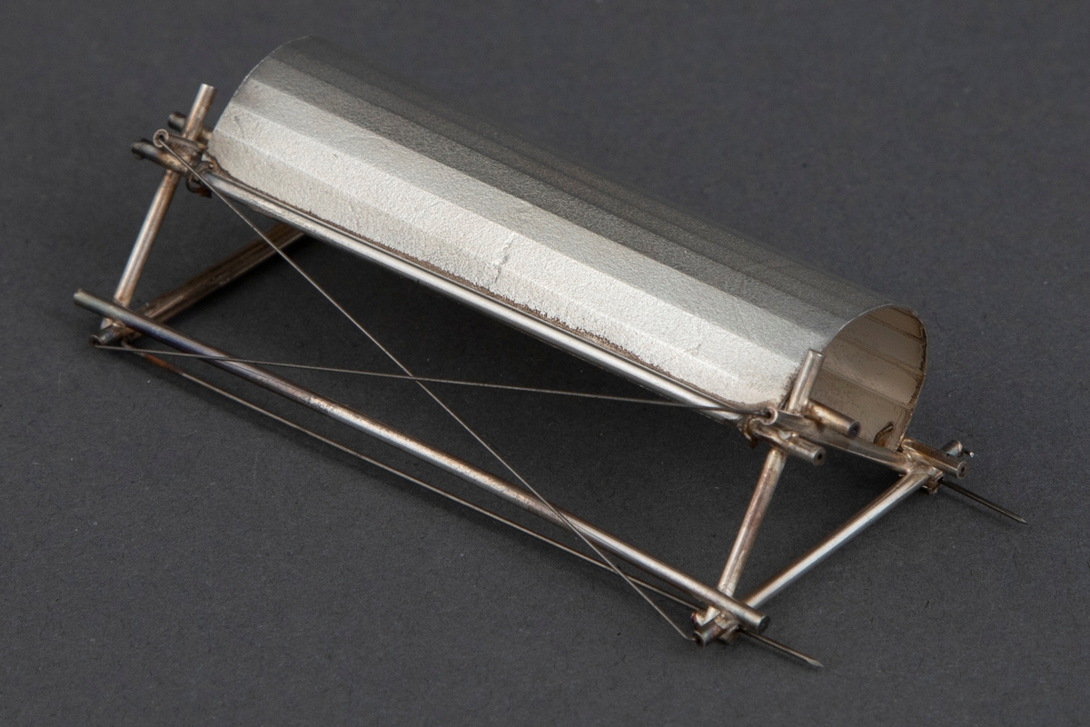 Brosje/smykkeobjekt formet av en rektangulær sølvplate som en halv sylinder med langsgående knekk. Denne er påsatt tynne sølvstrenger til en tilnærmet trekantform. Videre er det påsatt tynne ståltråder til dekor og som nåler til å feste brosjen.