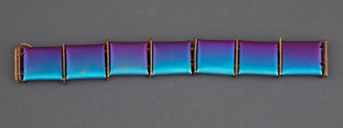 Leddet armbånd av titan. Leddene består av kvadratiske plater i magenta og blågrønt med en skimrende overflate. På baksiden er hjørnene brettet inn mot hverandre slik at leddene blir "pute"-formet. Kvadratene er tredd inn på to tynne metallkjettinger og med rektangulære gullfargede plater mellom hvert ledd. Låsen består av en metallpinne som stikkes ned i et rør.