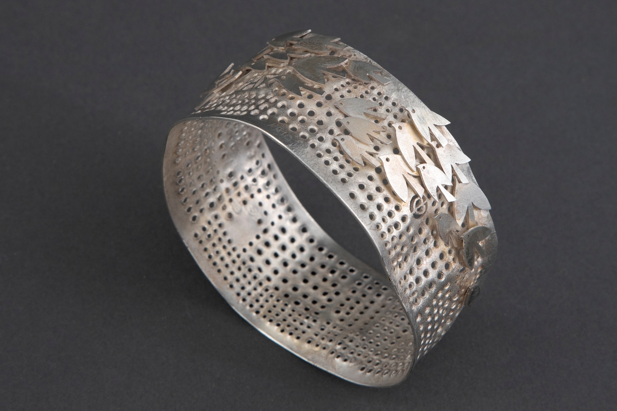Tilnærmet sirkulær armring i sølv. Overflaten er perforert, og dekorert stiliserte fugler.