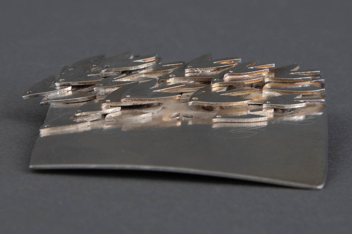 Tilnærmet kvadratisk brosje i sølv. Brosjens overside er dekorert med en rekke stiliserte duer i høyt relieff, som er loddet fast til overflaten. Konturene til fire duelignende fugler er gravert på brosjens nedre del. På baksiden er det gravert "KRIG ER FORAKT FOR LIV / FRED ER Å SKAPE / N. GRIEG", flankert av konturene av to duer. Påsatt nål.