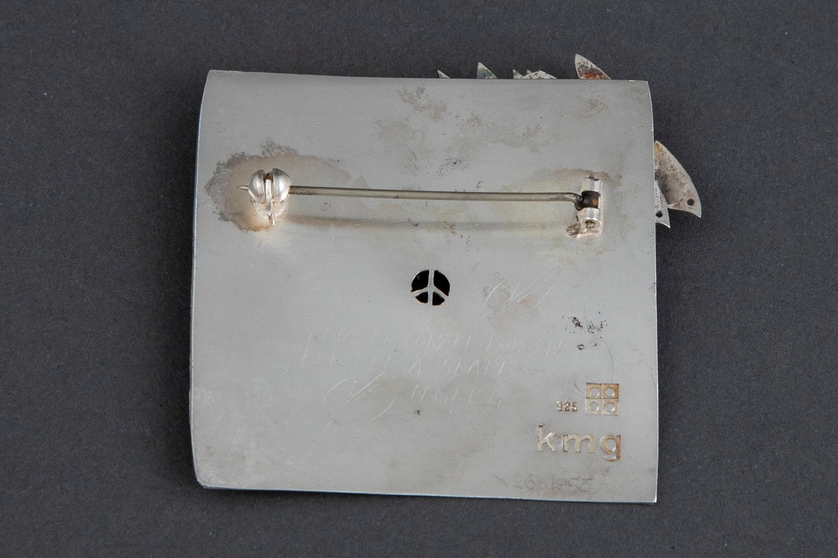 Tilnærmet kvadratisk brosje i sølv. Brosjens overside er dekorert med en rekke stiliserte duer i høyt relieff, som er loddet fast til overflaten. Konturene til fire duelignende fugler er gravert på brosjens nedre del. På baksiden er det gravert "KRIG ER FORAKT FOR LIV / FRED ER Å SKAPE / N. GRIEG", flankert av konturene av to duer. Påsatt nål.