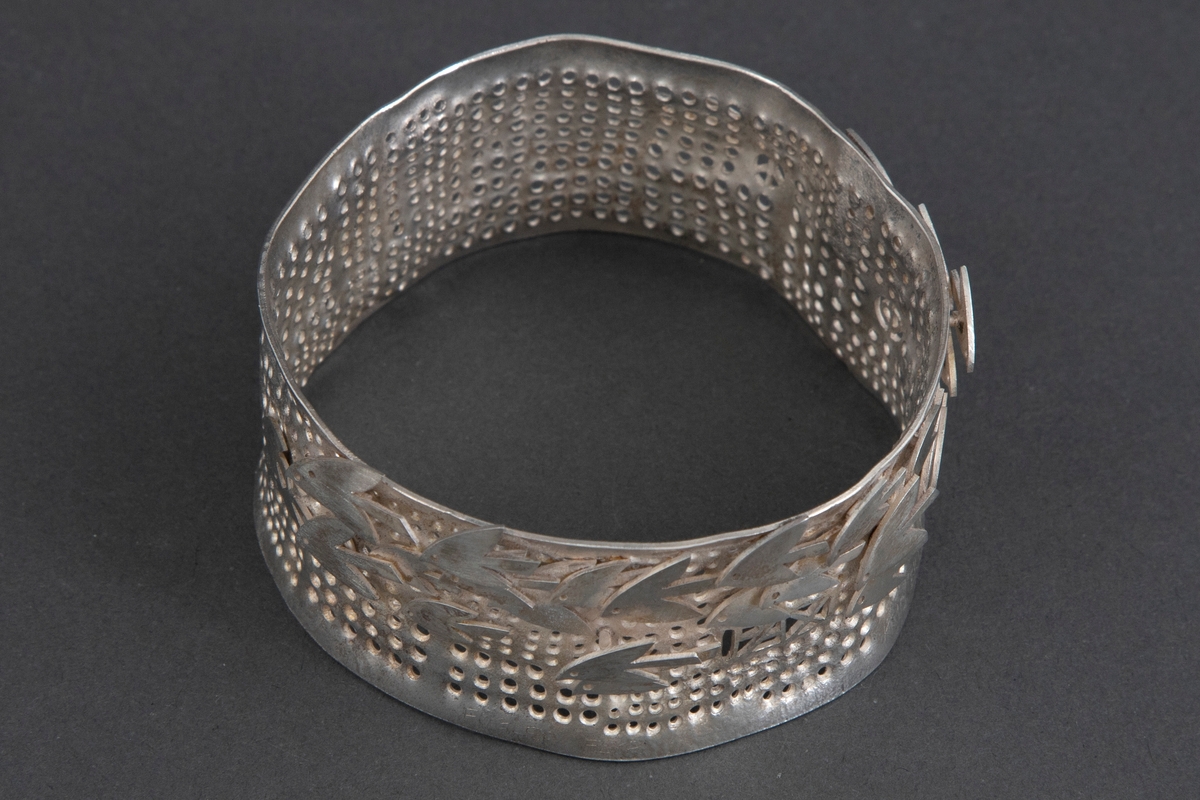 Tilnærmet sirkulær armring i sølv. Overflaten er perforert, og dekorert stiliserte fugler.