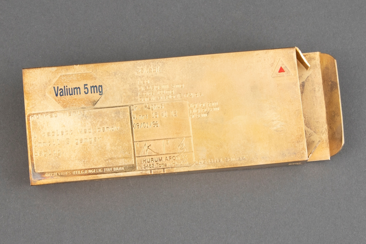 Brosje i rektangulær eskeform i forgylt bronse. Smykket er en kopi av en medisineske som er åpnet i den ene enden. Esken "inneholder" Valium 5mg utskrevet til Konrad Mehus av Dr. Magnus 09/01/96. Alle sider har tekst i opphøyd skrift: Innhold, fabrikkmerke osv. Brosjen har to lange nåler av stål på baksiden.
