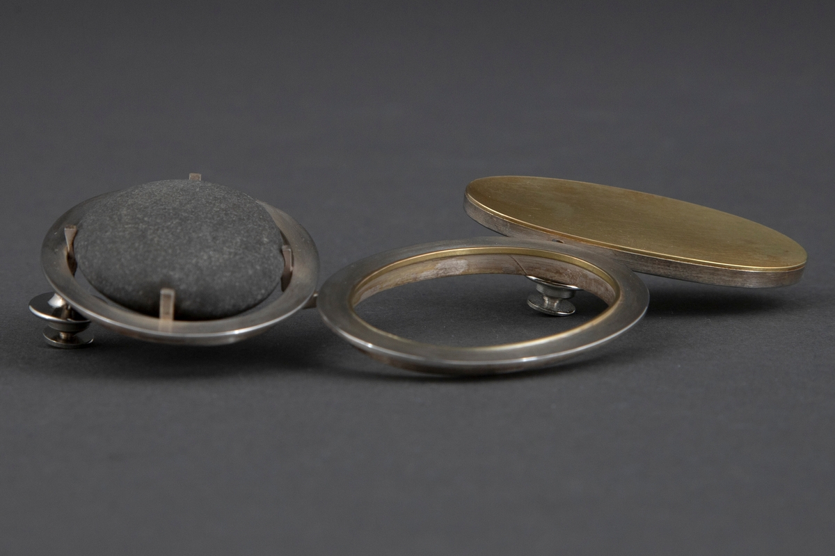 Brosjen er satt sammen av tre ovale ringer i børstet sølv og gull. Sølvringen holder på en svart rullestein. Åpen ring i midten av sølv med en liten gullkant i veggene. Ringen til høyre har en heldekkende gullplate med sølvvegg. De tre ringene er festet sammen på baksiden med sølvstaver. Brosjen har to pinfester. På baksiden av gullplaten er det inskribert signaturen Millie Behrens.