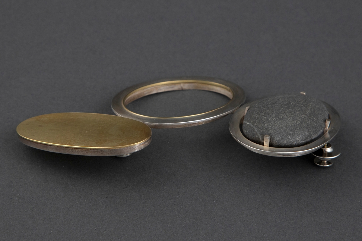 Brosjen er satt sammen av tre ovale ringer i børstet sølv og gull. Sølvringen holder på en svart rullestein. Åpen ring i midten av sølv med en liten gullkant i veggene. Ringen til høyre har en heldekkende gullplate med sølvvegg. De tre ringene er festet sammen på baksiden med sølvstaver. Brosjen har to pinfester. På baksiden av gullplaten er det inskribert signaturen Millie Behrens.
