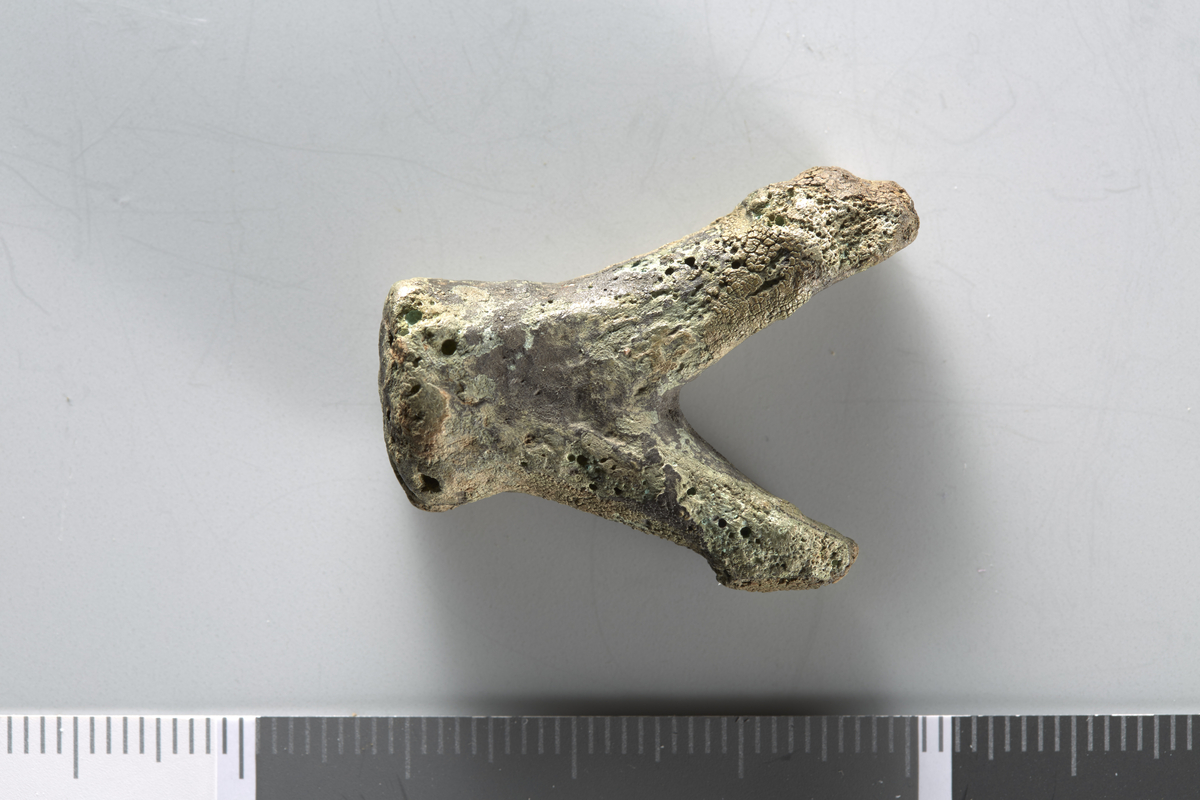 V-formet bronsefragment, muligens støpeknoll. Største lengde er 3,1 cm, bredden over "benene" er 2,5 cm.