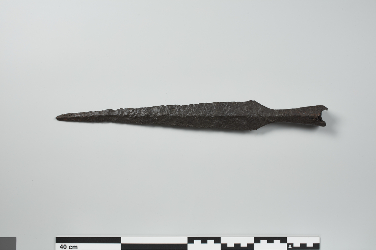 Stor spydspiss av jern. Av utseende minner den noe om R. 520, men avviker fra typeeksemplaret ved at den har fasetter på falen. Den er meget bra bevart. Bare et lite stykke nederst på falen er brukket av. Full lengde 40,7 cm, derav er falen 10,5 cm.