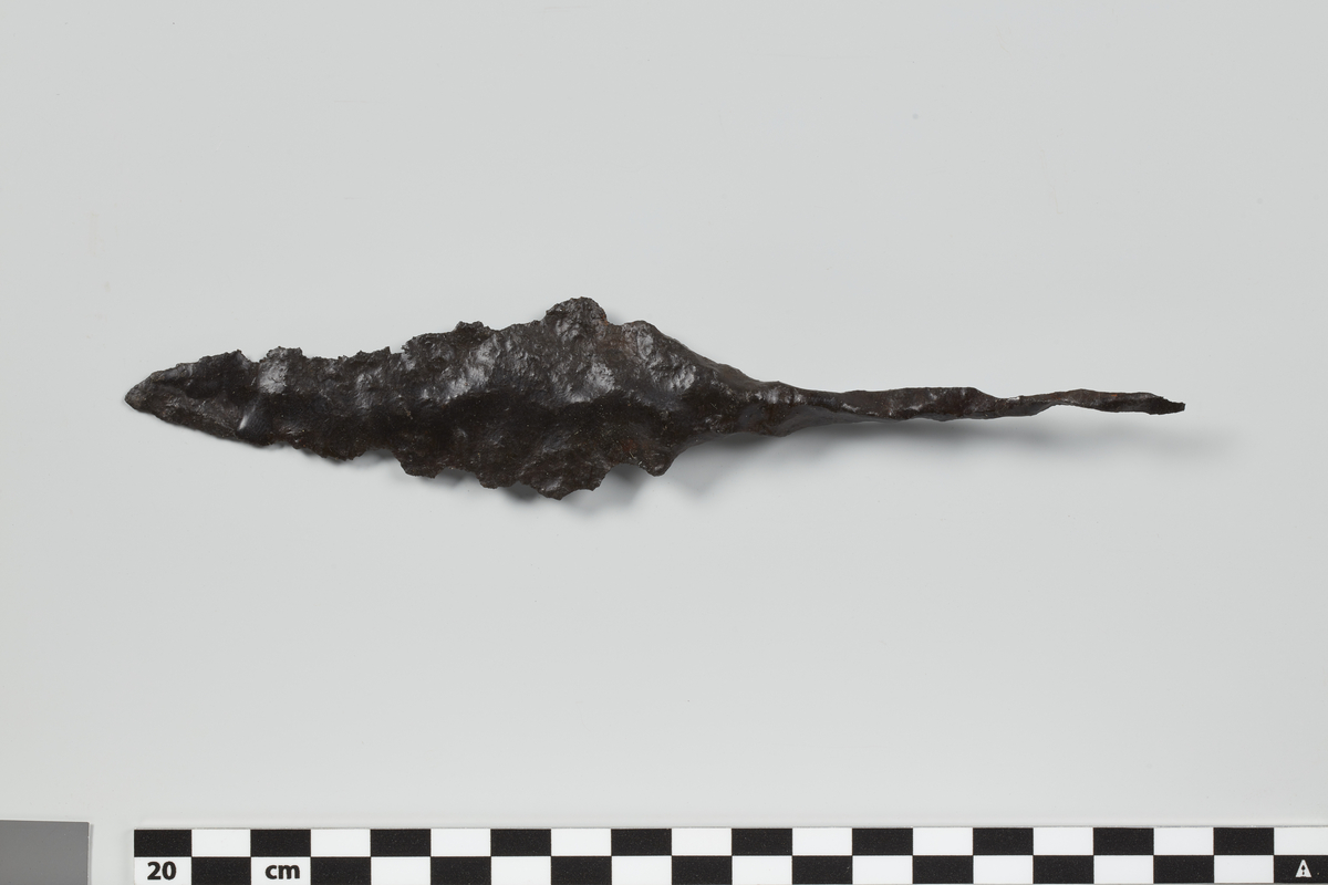 Liten kastespydspiss av jern av noe uvanlig form; harlang, slank tange, bladet nærmest formet som fig. 25 i Vikingesverd. En del medtatt. St. lengde 18 cm, bladets st. bredde 3,3 cm.