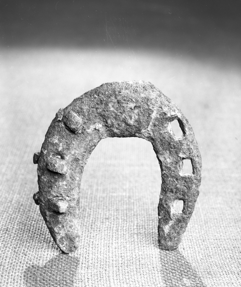 Hestesko av jern. Fortykket tåstykke. Ingen haker. 6 sømhull. 3 søm bevart. Sterkt forrustet. Lengde 10,0 cm, br. 9,5 cm.