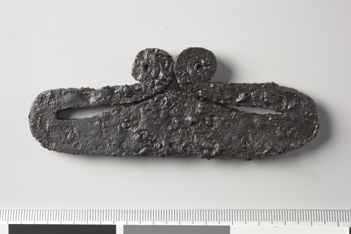 Ildstål av jern, R. 426, men atskillig større og med armene bøydd nesten helt inntil den breie tverrstang. Litt forrustet. Lengde 11,1 cm, største bredde 4,5 cm. 