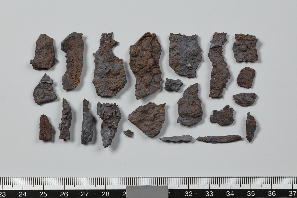 28 fragmenter av jern. 11 av stykkene er flate og kan stamme fra sverdet eller skjoldbulens krage. 1 stykke brent bein er korrodert fast på ett av fragmentene. De resterende 17 fragmentene består av svært små avskallinger. Stl. 3,3 cm.