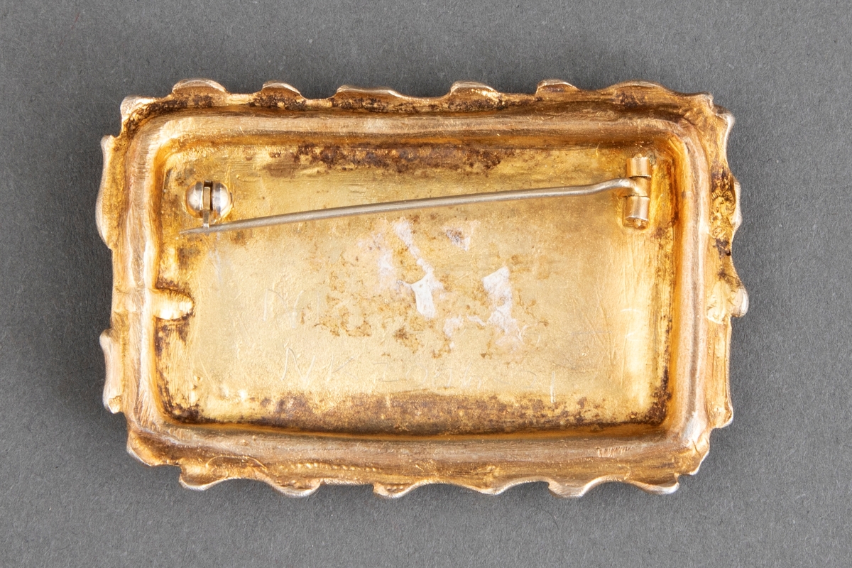 Rektangulær brosje i forgylt sølv med grønn emalje. Pastoralt motiv med fire stiliserte menneskefigurer og to dyr.