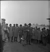 Skogsdag Örserums brunn 1961. Flera män står samlade på en lerig väg De flesta bär rock, slips och hatt och några bär gummistövlar. Två herrar står i mitten och pratar med varann. En av herrarna har en megafon hängande från armen.