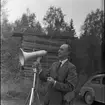 Skogsdag Örserums brunn 1961. En man står och håller upp en megafon framför ansiktet. Han håller en pipa i handen. Bakom honom finns en stapel med plank.