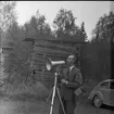 Skogsdag Örserums brunn 1961. En man står och håller upp en megafon framför ansiktet. Han håller en pipa i handen. Bakom honom finns en trave med plank.