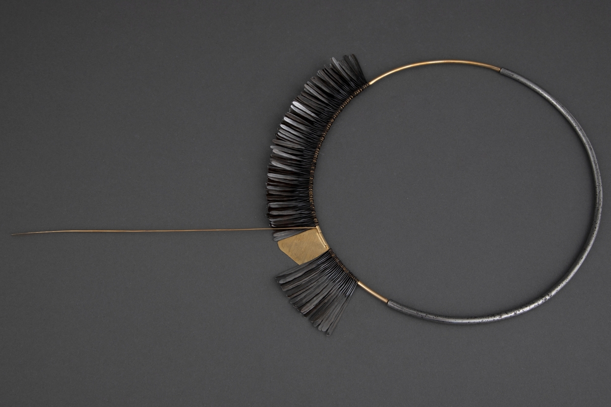 Halsklave bygget opp som en sirkel med en halvdel i gull og en halvdel i stål som er felt i hverandre. På 2/3 deler av gulldelen er det festet en krans av smidde spikre. Disse er kokt i linolje og har fått en mørk grå-svart farge. Litt nedenfor toppen av denne kransen er det festet en gullplate med skravert mønster. Fra denne løper en gullstav over og ned til den andre siden. Ved bruk løper denne foran halsen.