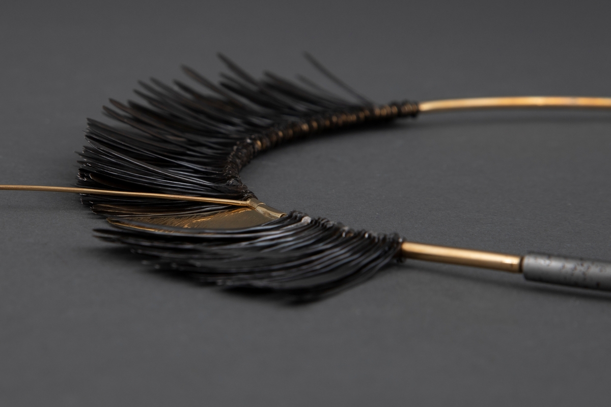 Halsklave bygget opp som en sirkel med en halvdel i gull og en halvdel i stål som er felt i hverandre. På 2/3 deler av gulldelen er det festet en krans av smidde spikre. Disse er kokt i linolje og har fått en mørk grå-svart farge. Litt nedenfor toppen av denne kransen er det festet en gullplate med skravert mønster. Fra denne løper en gullstav over og ned til den andre siden. Ved bruk løper denne foran halsen.