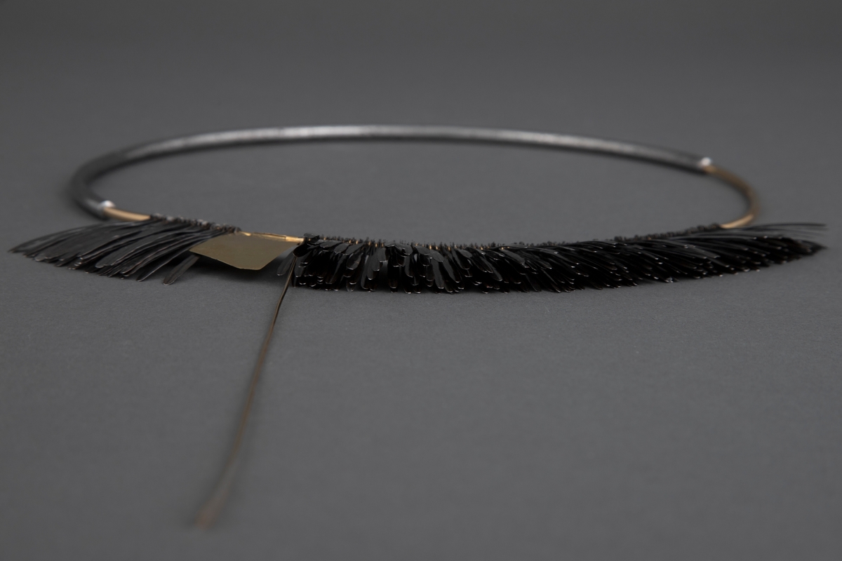 Halsklave bygget opp som en sirkel med en halvdel i gull og en halvdel i stål som er felt i hverandre. På 2/3 deler av gulldelen er det festet en krans av smidde spikre. Disse er kokt i linolje og har fått en mørk grå-svart farge. Litt nedenfor toppen av denne kransen er det festet en gullplate med skravert mønster. Fra denne løper en gullstav over og ned til den andre siden. Ved bruk løper denne foran halsen.