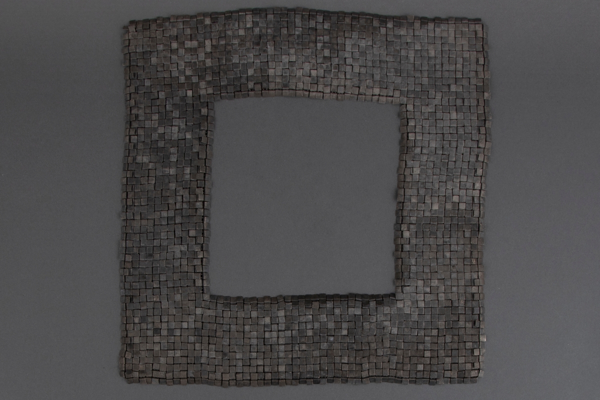 Kvadratisk smykke med kvadratisk halsåpning i oksydert sølv. Overflaten består av små kvadrater, som egentlig er rektangler som er brettet mot baksiden. Her er de hektet sammen med øskener, slik at fremsiden fremstår som en tett flate. Smykket trees over hodet.