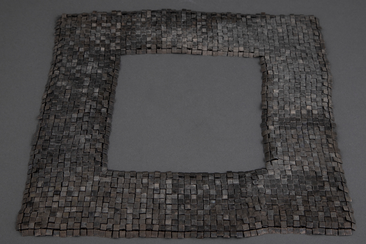 Kvadratisk smykke med kvadratisk halsåpning i oksydert sølv. Overflaten består av små kvadrater, som egentlig er rektangler som er brettet mot baksiden. Her er de hektet sammen med øskener, slik at fremsiden fremstår som en tett flate. Smykket trees over hodet.