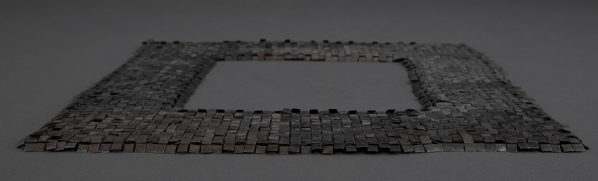 Kvadratisk smykke med kvadratisk halsåpning i oksydert sølv. Overflaten består av små kvadrater, som egentlig er rektangler som er brettet mot baksiden. Her er de hektet sammen med øskener, slik at fremsiden fremstår som en tett flate. Smykket trees over hodet.