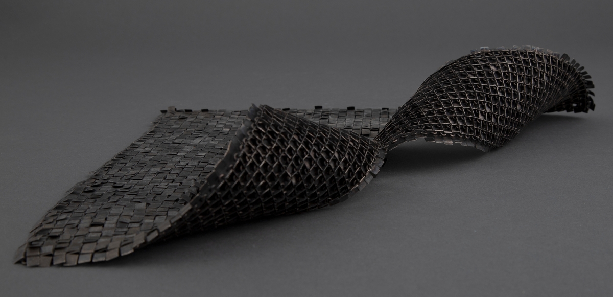 Kvadratisk smykke med kvadratisk halsåpning i oksydert sølv. Overflaten består av små kvadrater, som egentlig er rektangler som er brettet mot baksiden. Her er de hektet sammen med øskener, slik at fremsiden fremstår som en tett flate. Smykket trees over hodet.