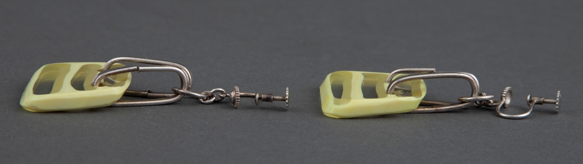 2) Ørepynt i par med stor, trapesformet glassbit i gult og klart glass, der klare og gule horisontale striper er omkranset av gult. Glassbiten er festet med to sølvringer til en mindre ring og har festeskrue for øre.