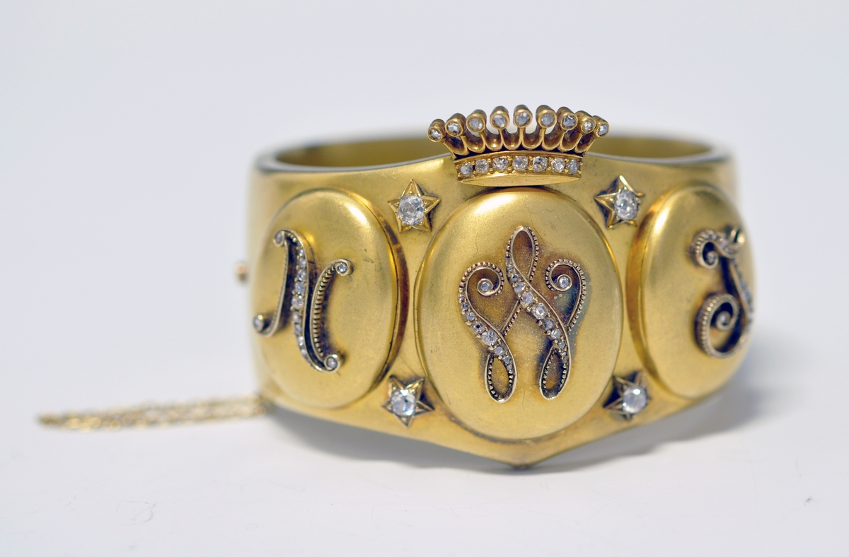 A.Bredt gullarmbånd som kan åpnes  på siden med lås og sikkerhetslenke. Armbåndet går i en spiss foran og det er montert en krone øverst men flere små diamanter, kronen er noe løs. Foran har armbåndet tre lokk  som kan åpnes. Framsiden av lokkene er dekoret med initialer i diamanter :  NWJ. Hvert lokk kan åpnes og inneholder fire fotografier av mannspersoner.Innvendig er det gravert inn årstall og stemplet.

B. Lilla eske smykkeeske som er tilpasset armbåndets form og har lås. Innvendig i lokket er firmaets navn stemplet.