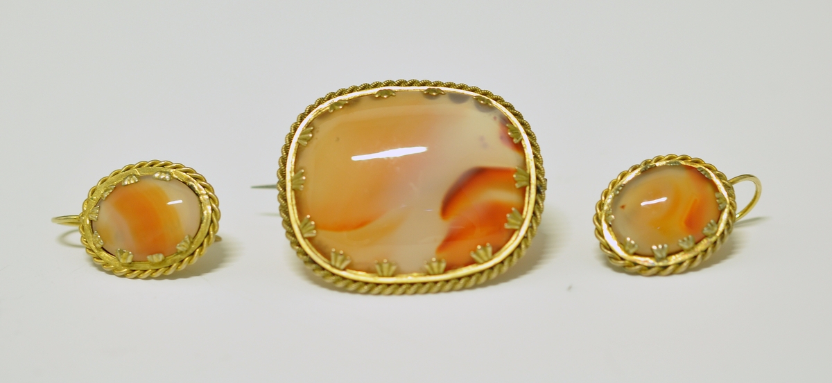 Smykkesett som består av 1 stk brosje, og 2 stk øreheng. A.Oval nesten kvadratisk brosje som består av  brun-orange stein (agatl?). Steinen er omkranset av smal metallkant  som er snodd rundt steinen, og  festet med små "blad" Steinen vises også på baksiden, og en smal nål er montert på.

B. og C. Øreheng/pynt i samme utførelse som brosjen, det er montert en lang bøyle til feste.  

D. Brunrød smykkeeske medfølger, på baksiden kan det være et stempel eller nr?