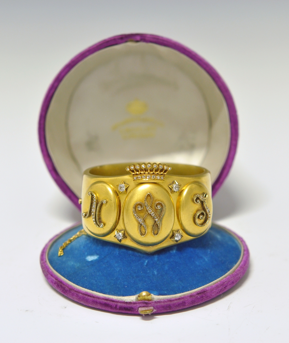 A.Bredt gullarmbånd som kan åpnes  på siden med lås og sikkerhetslenke. Armbåndet går i en spiss foran og det er montert en krone øverst men flere små diamanter, kronen er noe løs. Foran har armbåndet tre lokk  som kan åpnes. Framsiden av lokkene er dekoret med initialer i diamanter :  NWJ. Hvert lokk kan åpnes og inneholder fire fotografier av mannspersoner.Innvendig er det gravert inn årstall og stemplet.

B. Lilla eske smykkeeske som er tilpasset armbåndets form og har lås. Innvendig i lokket er firmaets navn stemplet.
