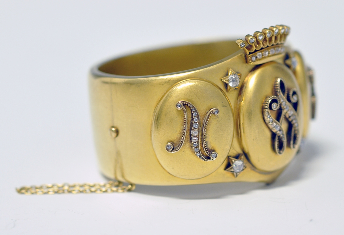 A.Bredt gullarmbånd som kan åpnes  på siden med lås og sikkerhetslenke. Armbåndet går i en spiss foran og det er montert en krone øverst men flere små diamanter, kronen er noe løs. Foran har armbåndet tre lokk  som kan åpnes. Framsiden av lokkene er dekoret med initialer i diamanter :  NWJ. Hvert lokk kan åpnes og inneholder fire fotografier av mannspersoner.Innvendig er det gravert inn årstall og stemplet.

B. Lilla eske smykkeeske som er tilpasset armbåndets form og har lås. Innvendig i lokket er firmaets navn stemplet.