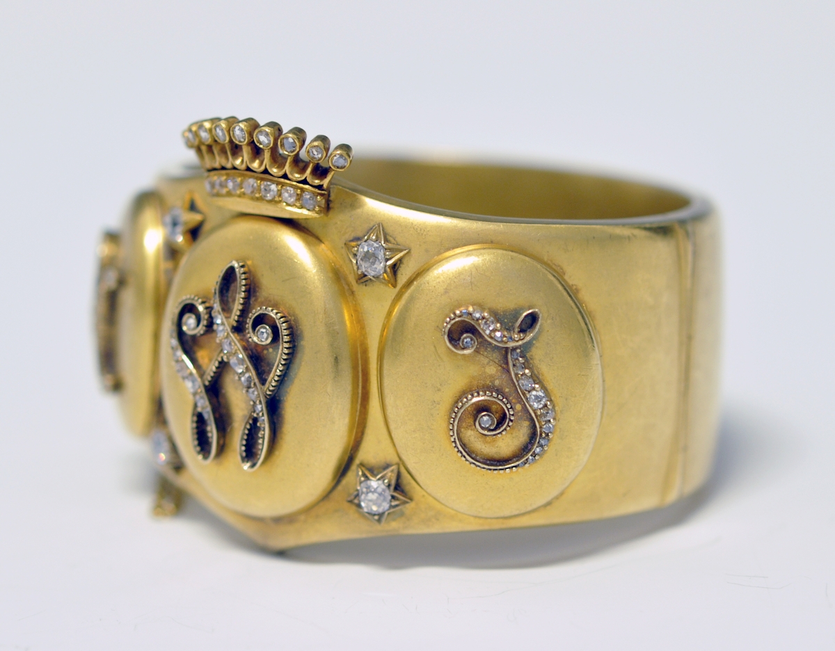 A.Bredt gullarmbånd som kan åpnes  på siden med lås og sikkerhetslenke. Armbåndet går i en spiss foran og det er montert en krone øverst men flere små diamanter, kronen er noe løs. Foran har armbåndet tre lokk  som kan åpnes. Framsiden av lokkene er dekoret med initialer i diamanter :  NWJ. Hvert lokk kan åpnes og inneholder fire fotografier av mannspersoner.Innvendig er det gravert inn årstall og stemplet.

B. Lilla eske smykkeeske som er tilpasset armbåndets form og har lås. Innvendig i lokket er firmaets navn stemplet.