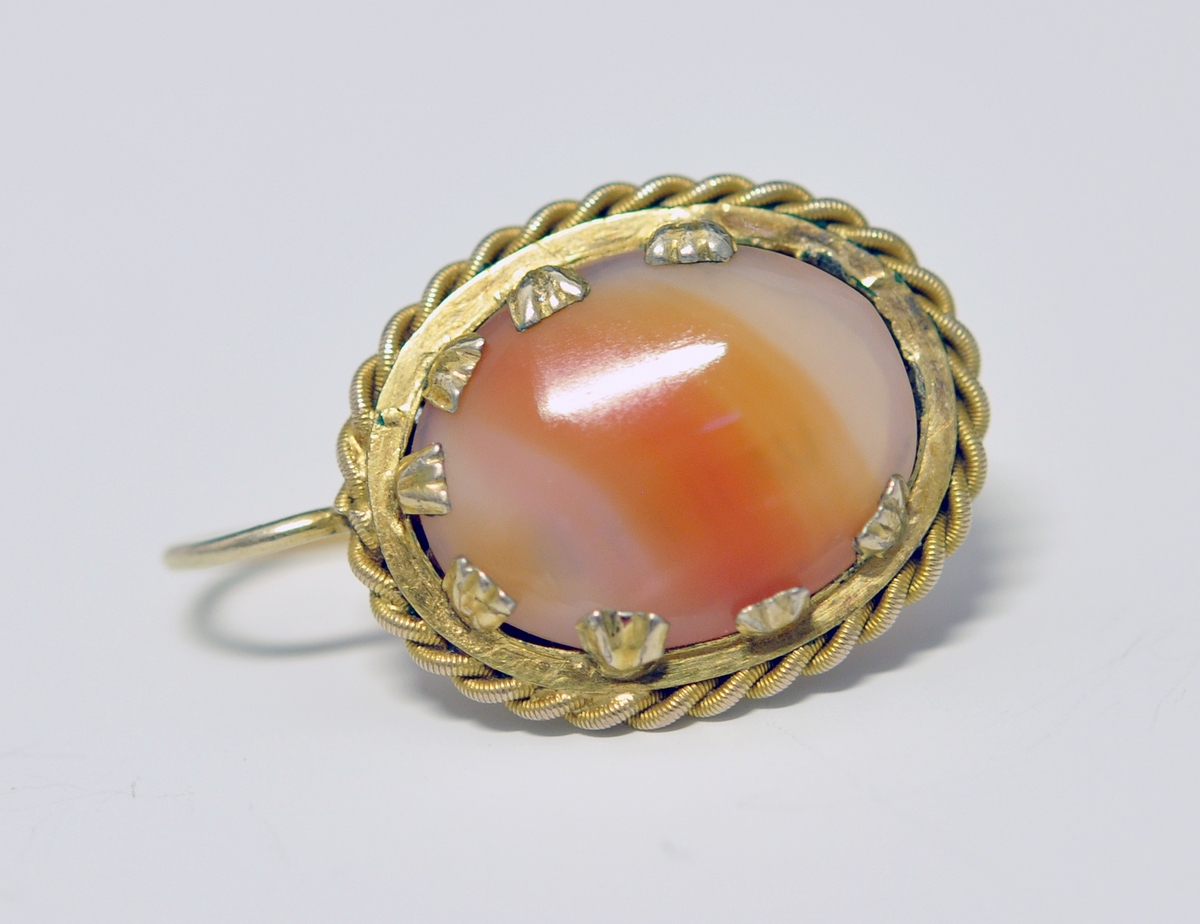 Smykkesett som består av 1 stk brosje, og 2 stk øreheng. A.Oval nesten kvadratisk brosje som består av  brun-orange stein (agatl?). Steinen er omkranset av smal metallkant  som er snodd rundt steinen, og  festet med små "blad" Steinen vises også på baksiden, og en smal nål er montert på.

B. og C. Øreheng/pynt i samme utførelse som brosjen, det er montert en lang bøyle til feste.  

D. Brunrød smykkeeske medfølger, på baksiden kan det være et stempel eller nr?