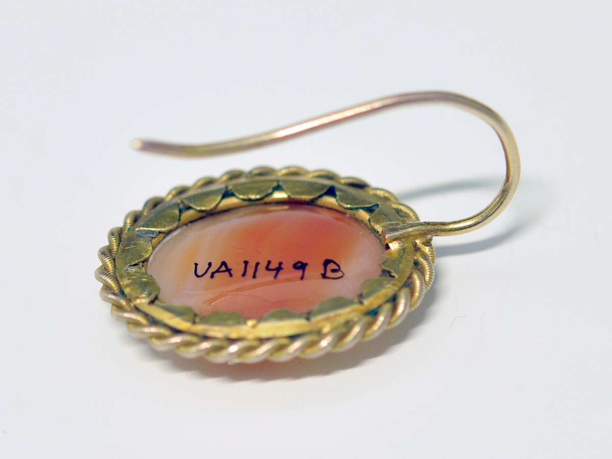 Smykkesett som består av 1 stk brosje, og 2 stk øreheng. A.Oval nesten kvadratisk brosje som består av  brun-orange stein (agatl?). Steinen er omkranset av smal metallkant  som er snodd rundt steinen, og  festet med små "blad" Steinen vises også på baksiden, og en smal nål er montert på.

B. og C. Øreheng/pynt i samme utførelse som brosjen, det er montert en lang bøyle til feste.  

D. Brunrød smykkeeske medfølger, på baksiden kan det være et stempel eller nr?
